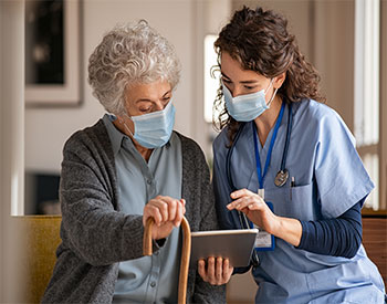 Doctor and senior woman going through medical record on digital tablet during home visit wearing face mask.